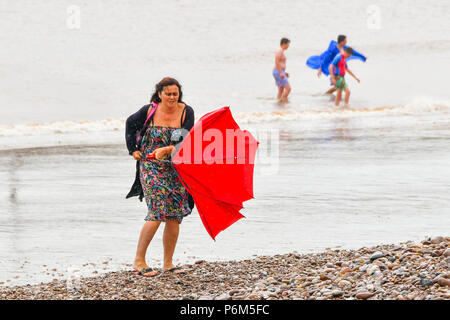 Sidmouth, Devon, UK.  1st July 2018. UK Weather.  A woman on the beach at the seaside resort of Sidmouth in Devon with umbrella which is caught by a gust of wind which turns it inside out as the town catches the edge of a passing heavy rain shower.  Picture Credit: Graham Hunt/Alamy Live News Stock Photo