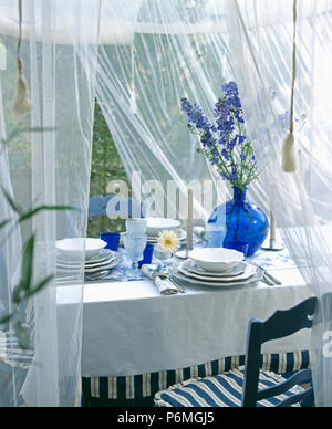 White voile drapes above dining table with set for lunch with white cloth and plates and blue delphiniums in blue vase Stock Photo