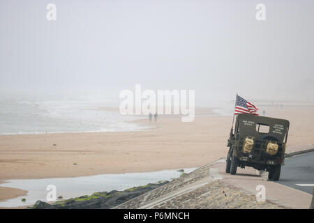 Vierville-sur-Mer, France. A Willys Jeep flying the US flag sits on the seawall overlooking Omaha Beach, as a distant couple walk along the waters edge on the anniversary of D Day Stock Photo