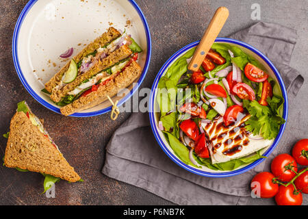 Bright summer salad with tomatoes, onions, balsamic and goat cheese with meat and cheese sandwiches. Food flat lay on dark background. Stock Photo