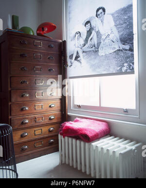 Black+white photograph-patterned blind on window above large radiator in study with tall antique chest-of-drawers Stock Photo