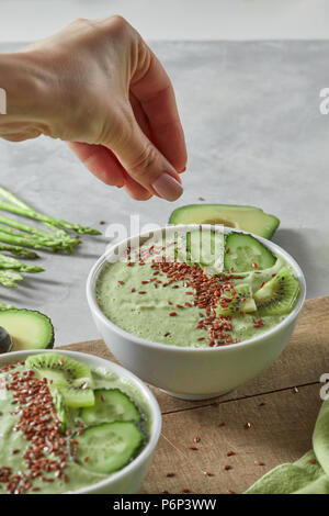 A girl's hand is laying flax seeds in a plate with fresh vegetable smoothies from kiwi, cucumber and asparagus on a wooden board. Healthy diet food Stock Photo