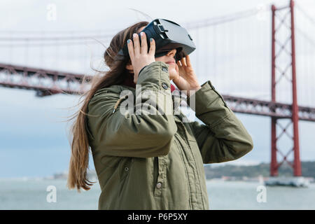 Young beautiful girl wearing virtual reality glasses. 25th of April bridge in Lisbon in the background. The concept of modern technologies and their use in everyday life Stock Photo