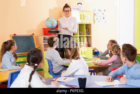 Cheerful female teacher giving geography lesson in classroom, showing pupils globe Stock Photo