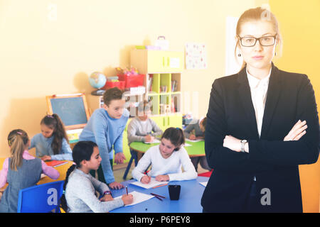 Strict diligent pleasant female teacher standing foreground in classroom with schoolchildren studying Stock Photo