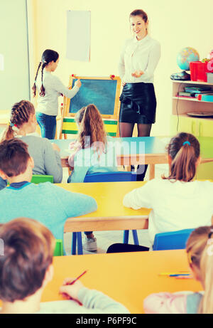 Positive friendly  diligent  female teacher standing at blackboard with schoolgirl during lesson in classroom Stock Photo