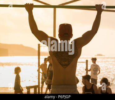 Rear view of muscular man with back tattoo doing pull ups/chin ups on beach in Spain Stock Photo