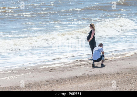 Skegness, UK. 2nd July 2018. A young couple on the beach at Skegness enjoying the current heatwave of hot weather, and temperatures as high as 30 degrees. The man is taking a photo of his pregnant partner while she stands on the beach with the waves at her feet. Credit: Steven Booth/Alamy Live News Stock Photo