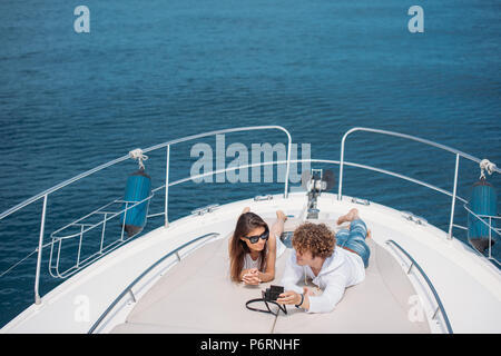 Attracrive young long-haired woman sitting on deck of luxury yacht being photographed by her curly handsome boyfriend over amazing seaviews with a lit Stock Photo