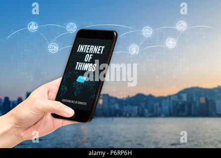 Hand holding mobile with Internet of things (iot) word on screen with icon feature glowing in air with blur city building background,Digital Lifestyle Stock Photo