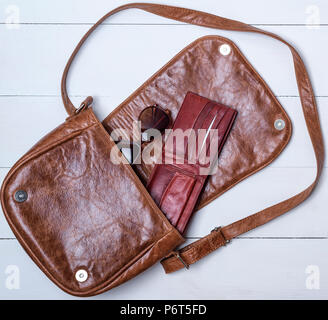 open brown leather bag with purse and glasses on a white wooden background, top view Stock Photo