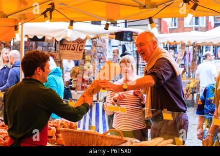 French bread being sold on market stall during Sandwich town's Le Weekend, French festival. Mature man reaching to take wrapped French stick loaf. Stock Photo