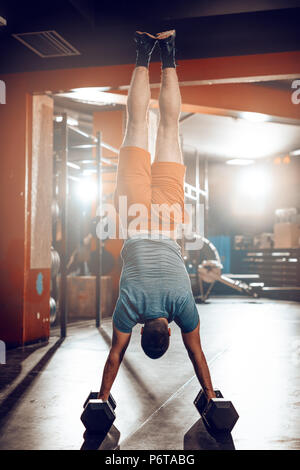 Young muscular man doing hard exercise at the gym. His is doing handstand and whole weight is on dumbbells. Stock Photo