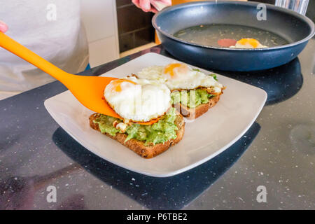 a fried egg being taken out of a frying pan on an orange plastic spatula and placed on avacado and brown toast on a plate Stock Photo