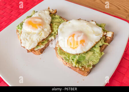 two fried eggs on avacado guacamole and brown toast on a white square shape plate, red place mat and a brown wood table Stock Photo