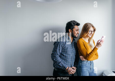 Satisfied happy mixed-race couple looking through pictures in social media while standing over grey wall background Stock Photo
