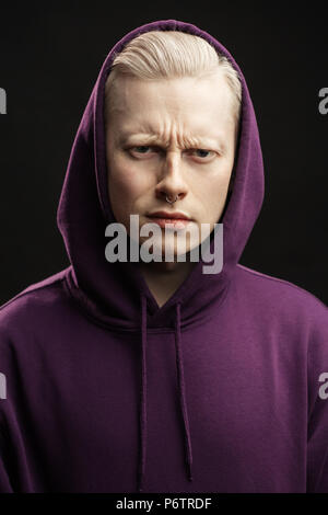 Blonde displeased guy in purple sweatshirt with a hood over his head frowning brows looking at camera over black background. Stock Photo