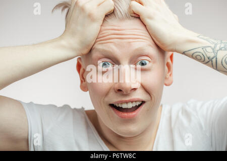 Close up headshot of overjoyed Albino hipster guy with bugged eyes and open mouth grabbing his hair in disbelief and happyness with tattooed hands. Hu Stock Photo