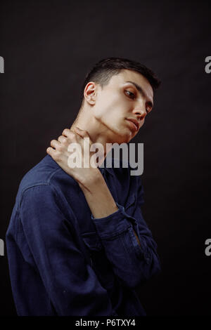 Good-looking upset brown haired young man looking as if he has neck problems or neck pain touching his neck, isolated, over black background Stock Photo