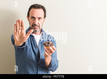 Senior man eating chocolate donut with open hand doing stop sign with serious and confident expression, defense gesture Stock Photo