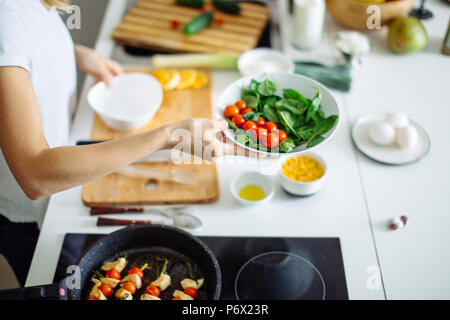 cropped view of woman hands making healthy salad from fresh vegetables, holding plate with tomato cherry and spinach. Frying pan with roasted chicken  Stock Photo