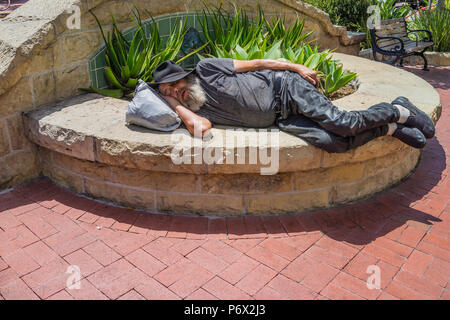 A bearded older male in a black fedora hat, grey t-shirt, black slacks and shoes, lies in a prone position sleeping on a city owned stone flower plant Stock Photo