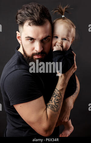 Studio portrait of loving young man hugging tight his adorable infant baby girl and looking at camera over black background. Stock Photo