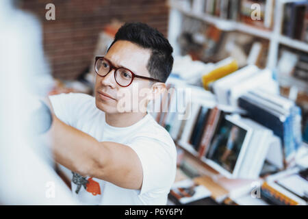 Young atrractive spectacled man drawing hand to book on a book shelf. Stock Photo
