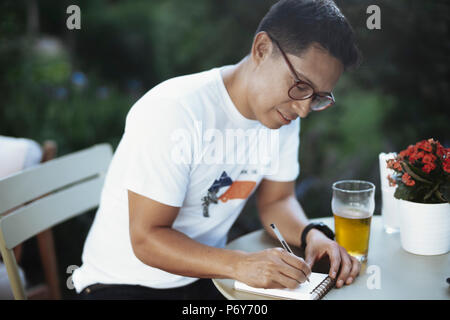 Young spectacled man drinking cold beer and writing some notes. Stock Photo