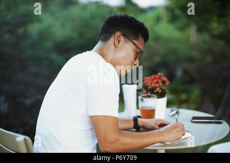 Young spectacled man drinking cold beer and writing some notes in notebook. Stock Photo
