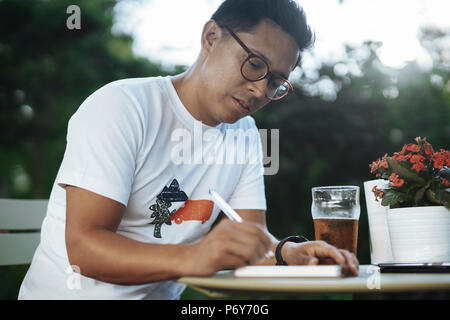 Young spectacled man drinking cold beer and writing some notes in notebook. Stock Photo