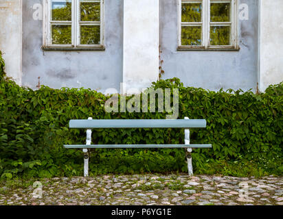 Old blue wooden bench in front of green shrubs and weathered blue house with two windows Stock Photo