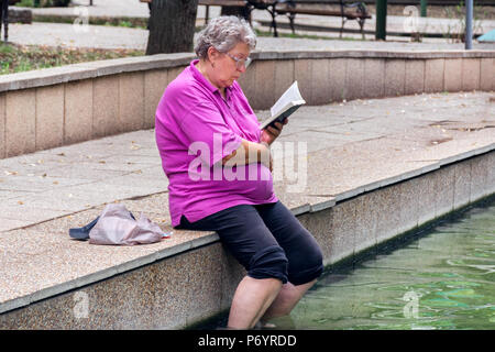 Niska Banja, Serbia - July 02, 2018: Senior lady reading book with legs in hot spa healthy water. Vacation and enjoyment of the summer concept Stock Photo