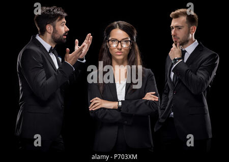confident businesswoman looking at camera while her colleagues discussing something behind, isolated on black Stock Photo