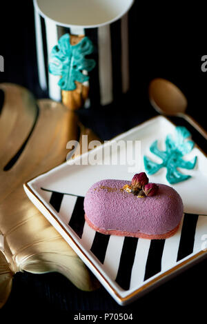 Purple fruit mousse cake, on a beautiful saucer.On a dark background, with a sheet of monstera painted in gold paint. Culinary art Stock Photo