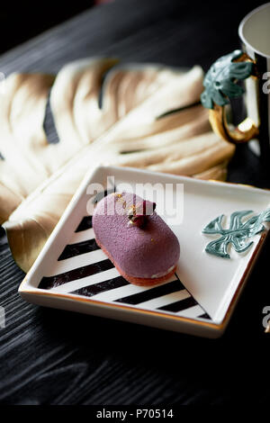 Purple fruit mousse cake, on a beautiful saucer.On a dark background, with a sheet of monstera painted in gold paint. Culinary art Stock Photo