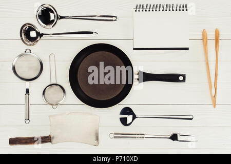top view of blank textbook, frying pan, butcher axe and kitchen utensils on white wooden table Stock Photo