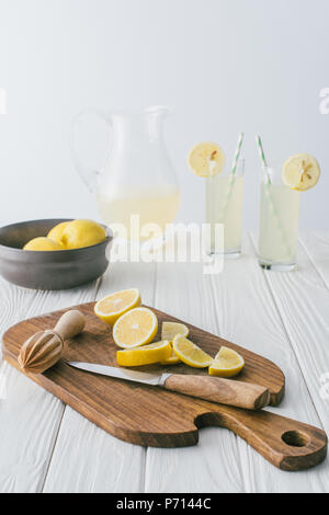 close up view of lemons, knife, wooden pestle on cutting board and lemonade in glasses on white wooden tabletop on grey backdrop Stock Photo