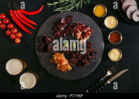 gourmet assorted meat on slate board, various sauces, vegetables and glasses of beer on black Stock Photo