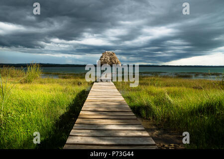 Dramatic clouds behind hut on a dock along the lake shore with dark blue cloudscape and sunshine, El Remate, Peten, Guatemala Stock Photo