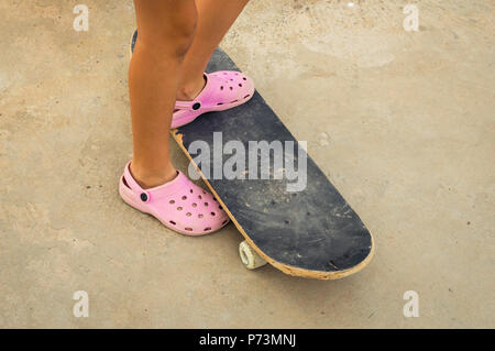 Little girl wearing pink plastic clogs riding on a skateboard. Learning to skate. Stock Photo