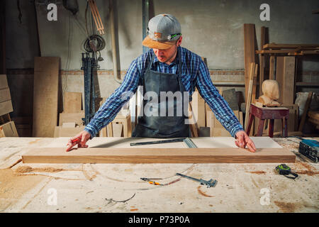 experienced carpenter in work clothes and small buiness owner measures a wooden board with a ruler and marks with pencil the necessary points for slic Stock Photo