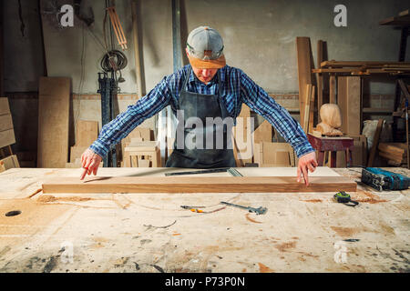 experienced carpenter in work clothes and small buiness owner measures a wooden board with a ruler and marks with pencil the necessary points for slic Stock Photo