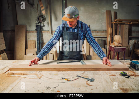 experienced carpenter in work clothes and small buiness owner measures a wooden board with a ruler and marks with pencil the necessary points for slic Stock Photo