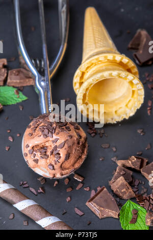 Chocolate ice cream in scoop with wafer sticks, cone and chocolate on a black slate board. Focus on ice cream in scoop. Stock Photo
