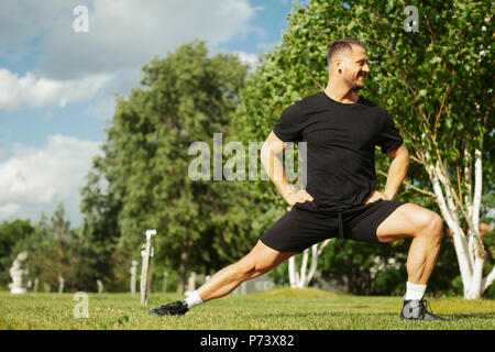 Young attractive man in black sportwear doing lunge outdoor in the park. Stock Photo