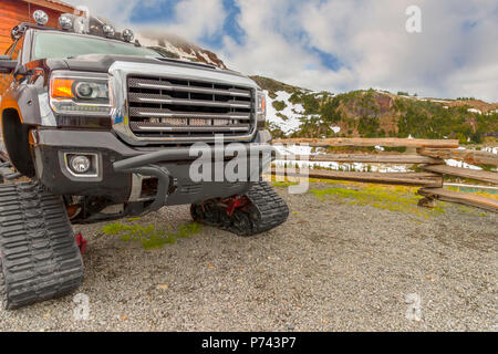 Car is on caterpillar tracks, stands near a wooden wall in snow-capped mountains, near a log fence, on a summer day with a blue sky and white white cl Stock Photo