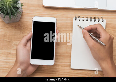 Overhead view of businessman hand holding smart phone while writing on notepad by camera and coffee on table Stock Photo