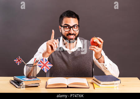 Handsome man in gray vest and eyeglasses has idea, holding red apple in hand, indoor shot at the office desk before black wall Stock Photo