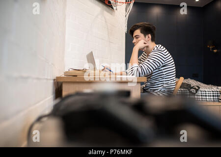 Attractive young male sitting at home at desk with books holding pen and using laptop computer. Stock Photo